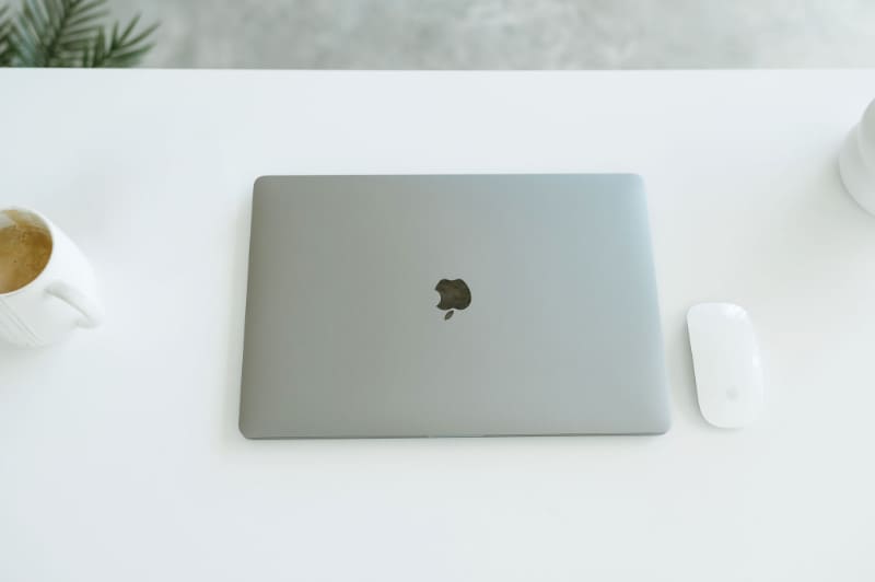 Closed MacBook on white desk with Apple mouse and coffee mug nearby. Clean setup for managing client systems and HoneyBook updates.