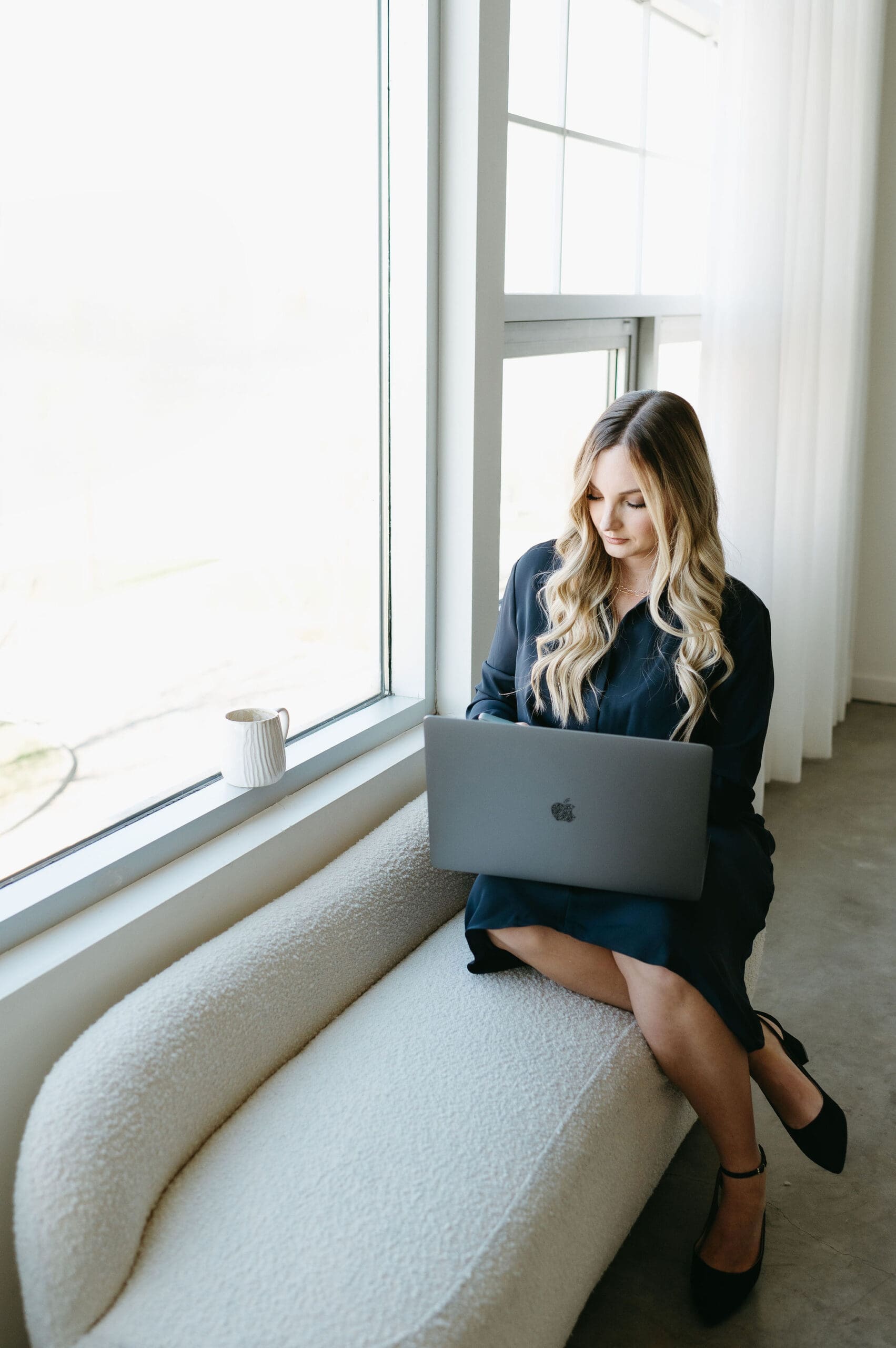Woman working on a laptop by a bright window, seated on a cozy cream bench with a coffee mug nearby. certified honeybok pros
