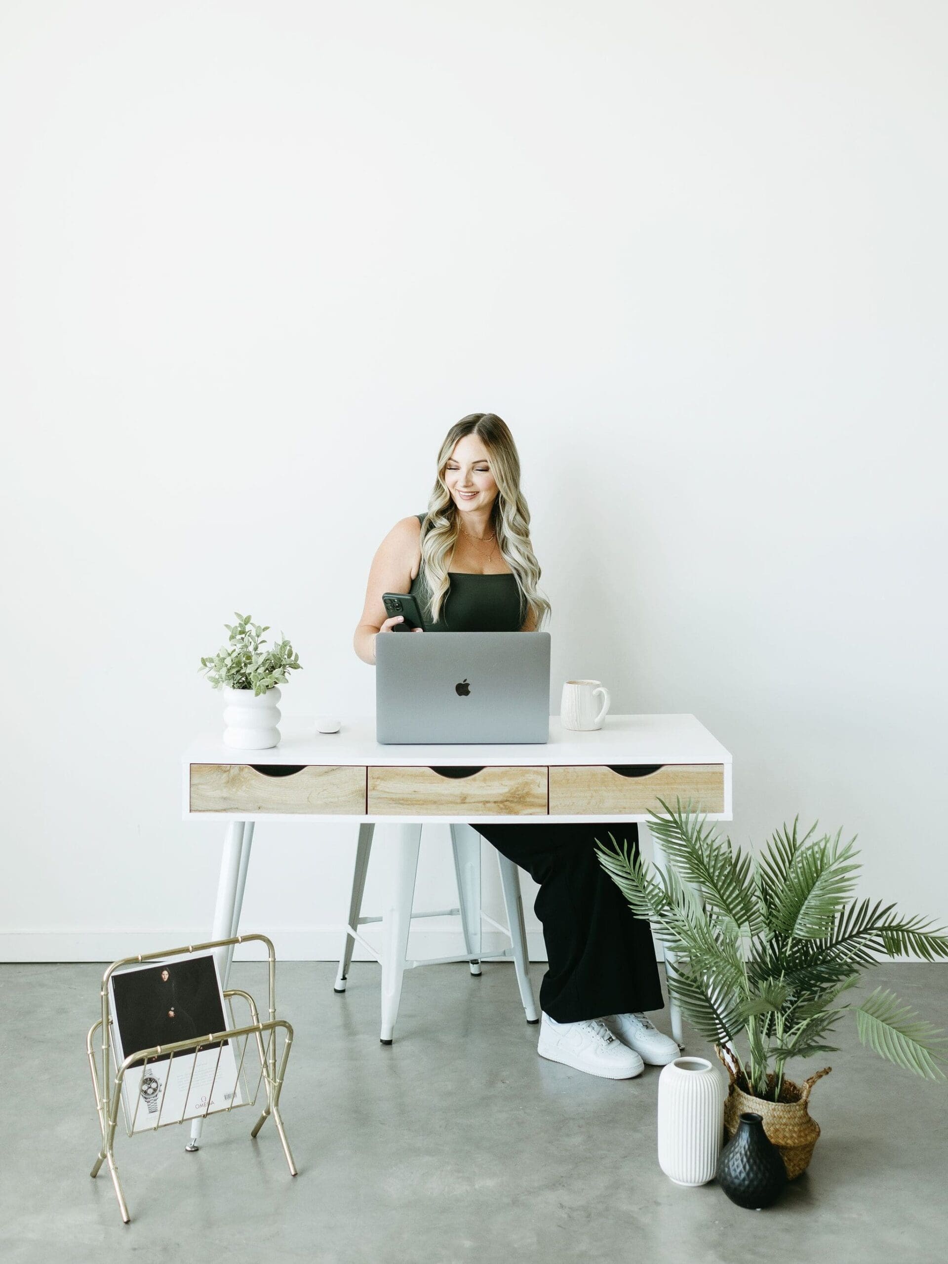 Woman smiling at her phone while working on a laptop at a modern white desk with plants and neutral decor.