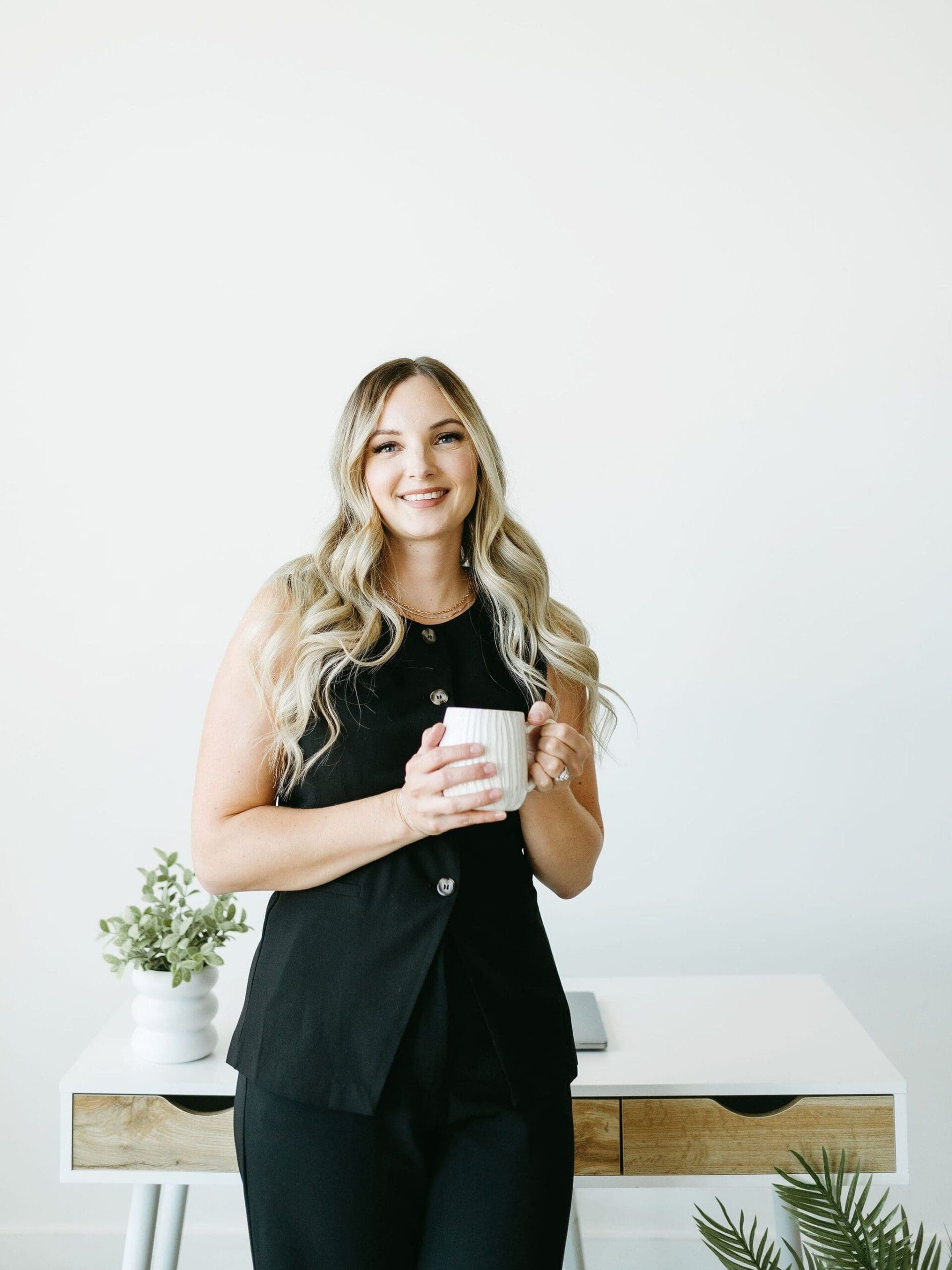 Casual, confident portrait of a woman holding a coffee mug, smiling in front of a desk with greenery and a light-filled background. certified honeybook pros
