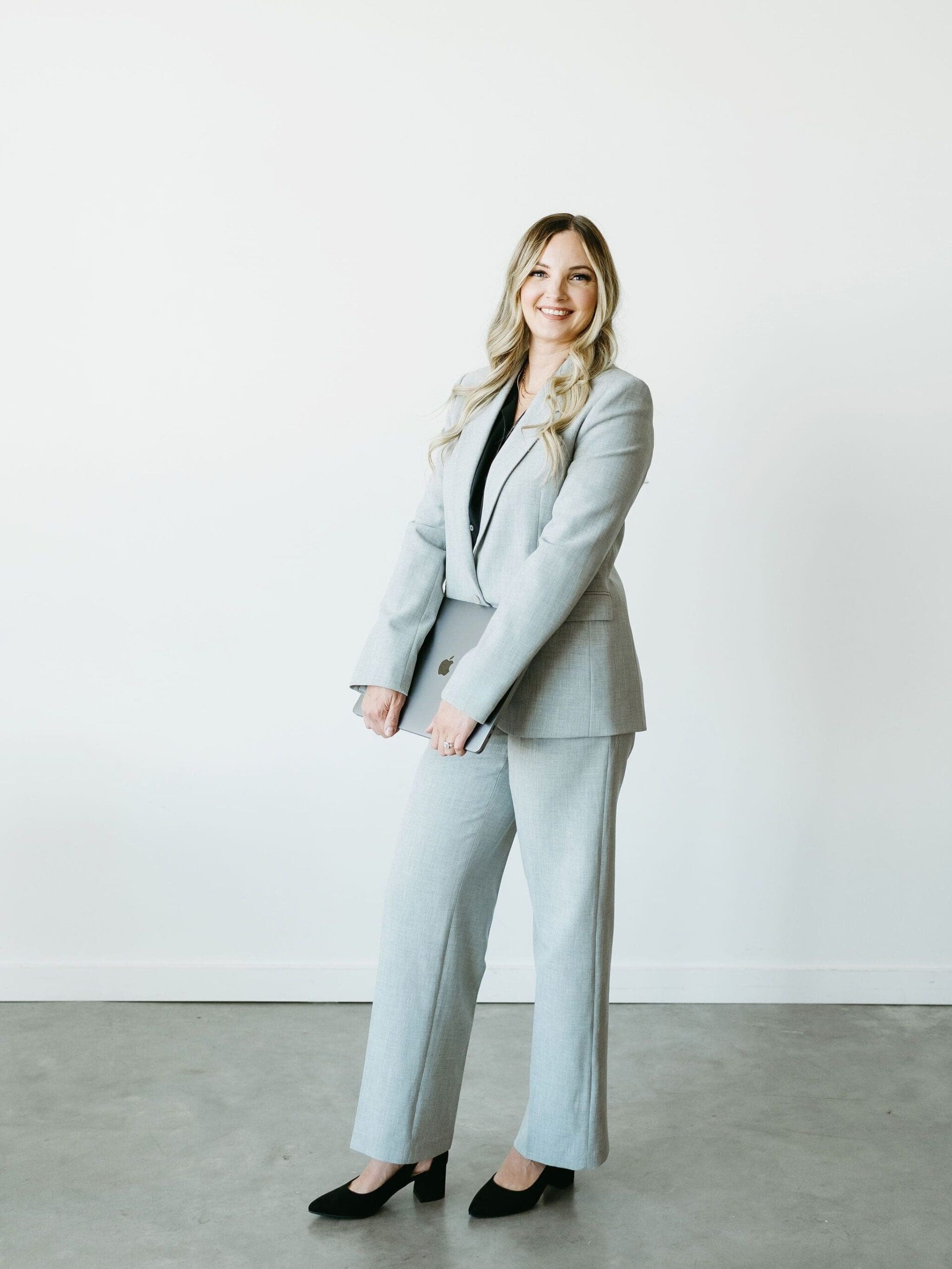 Professional woman in a light gray suit smiling confidently while holding a laptop against a clean, minimalist backdrop.