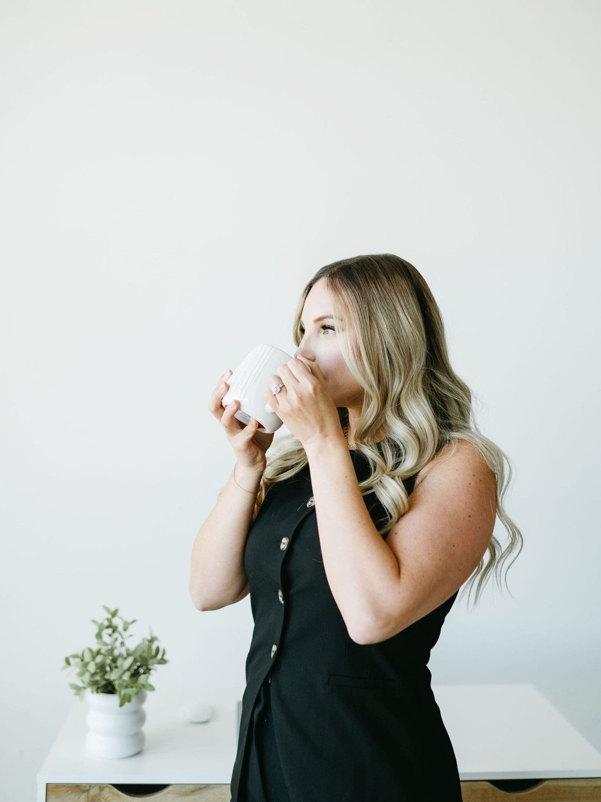 Woman sipping from a white mug while standing at her desk with relaxed posture, long waves, and a calm morning workspace feel.