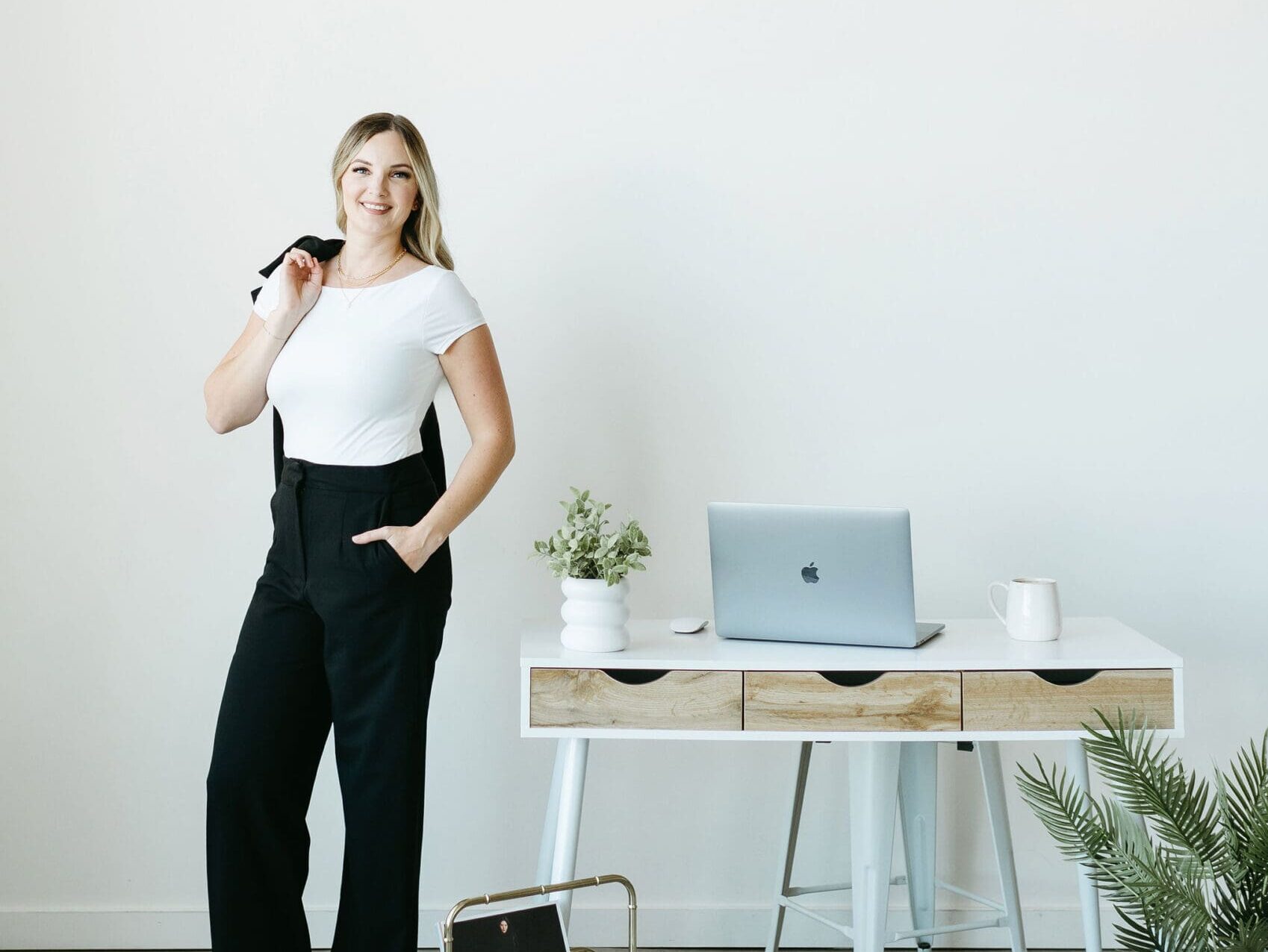 Woman standing confidently beside a minimalist desk with a laptop, plant, and mug, showcasing a clean, modern workspace for service providers. systems for service prodivers