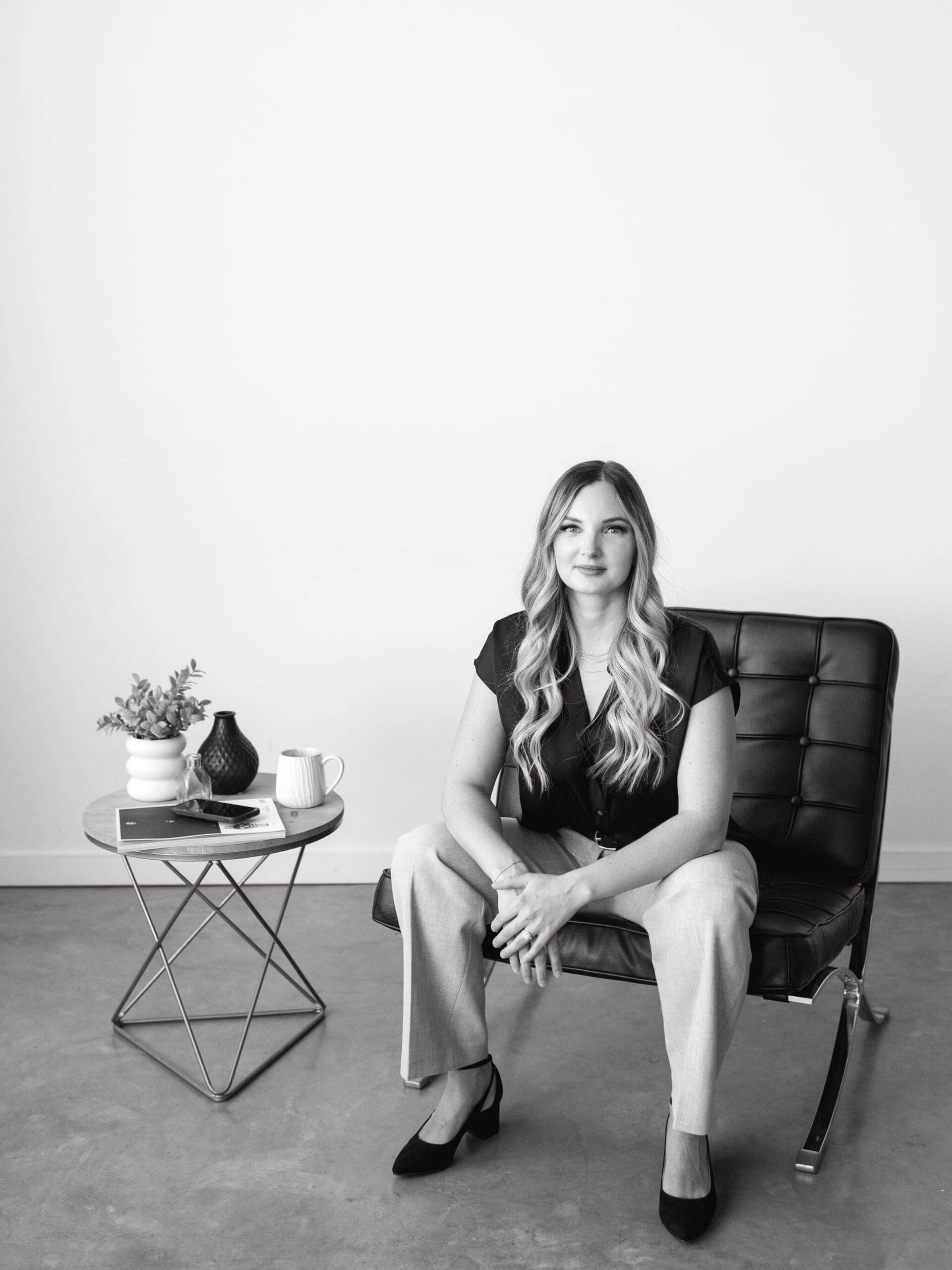 Woman seated in a modern chair with legs crossed, looking focused beside a small side table with coffee, greenery, and a phone — professional and approachable vibe. systems for service providers