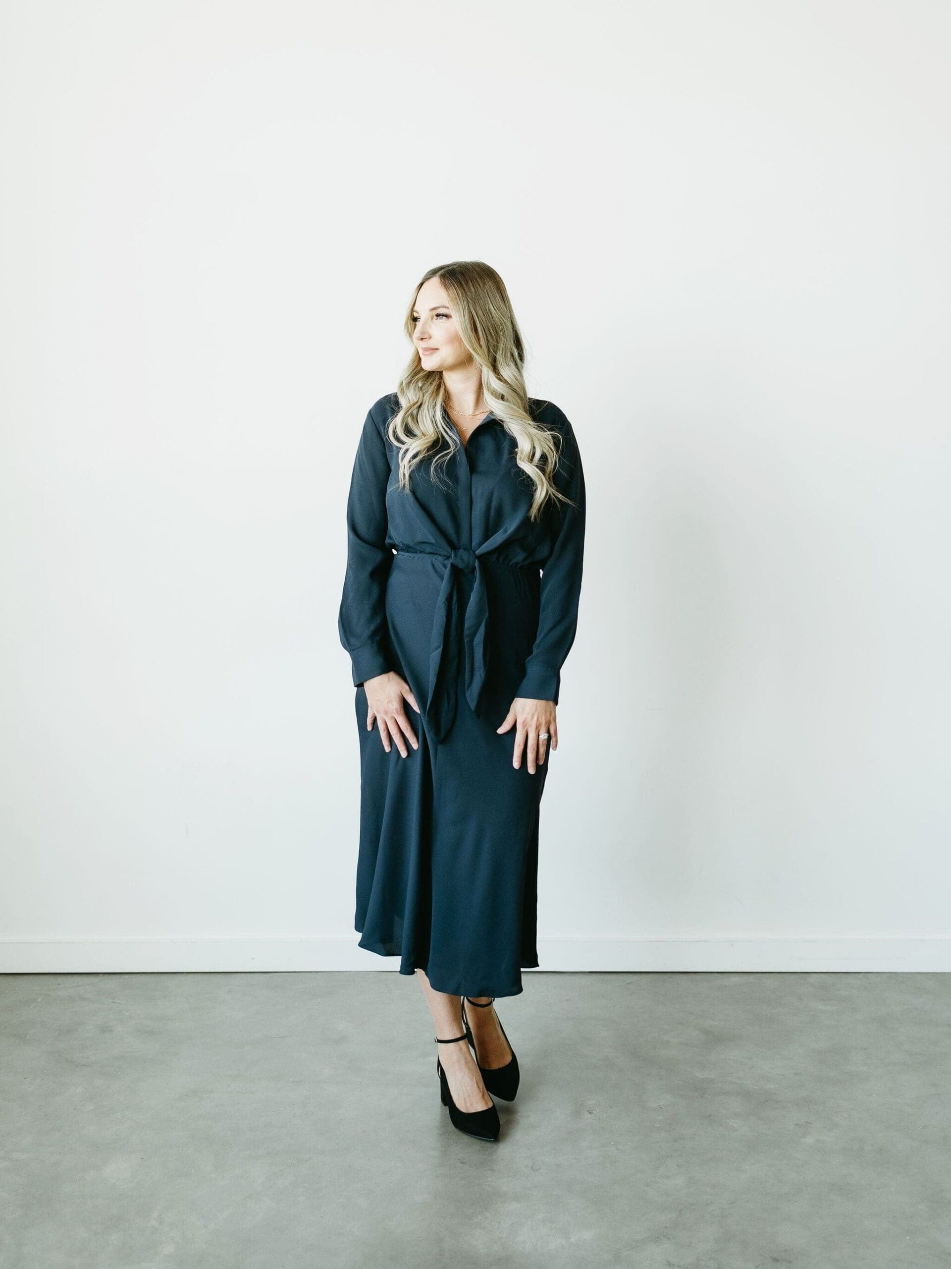 Woman in a navy dress standing in a bright, minimalist room, looking to the side with a soft smile — polished, poised, and client-ready.