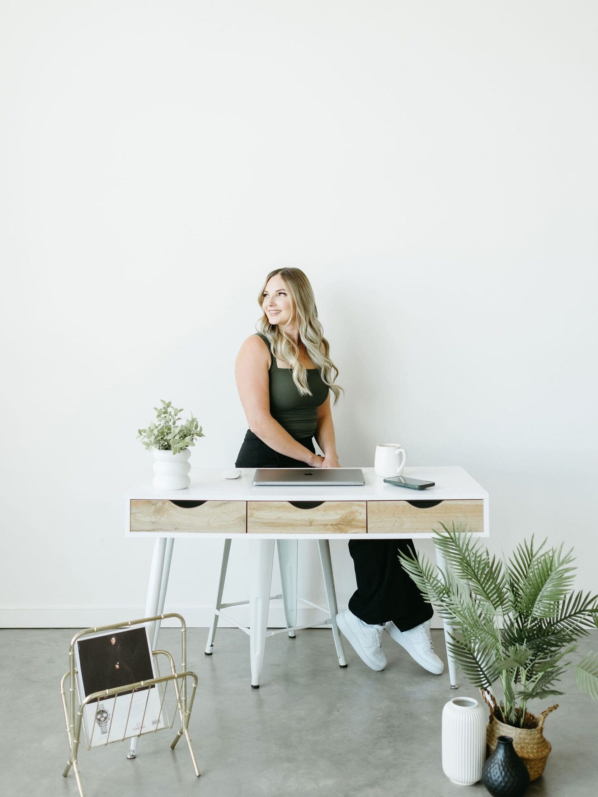 Woman standing behind a modern white desk with a laptop, phone, coffee mug, and plants in a bright minimalist office.