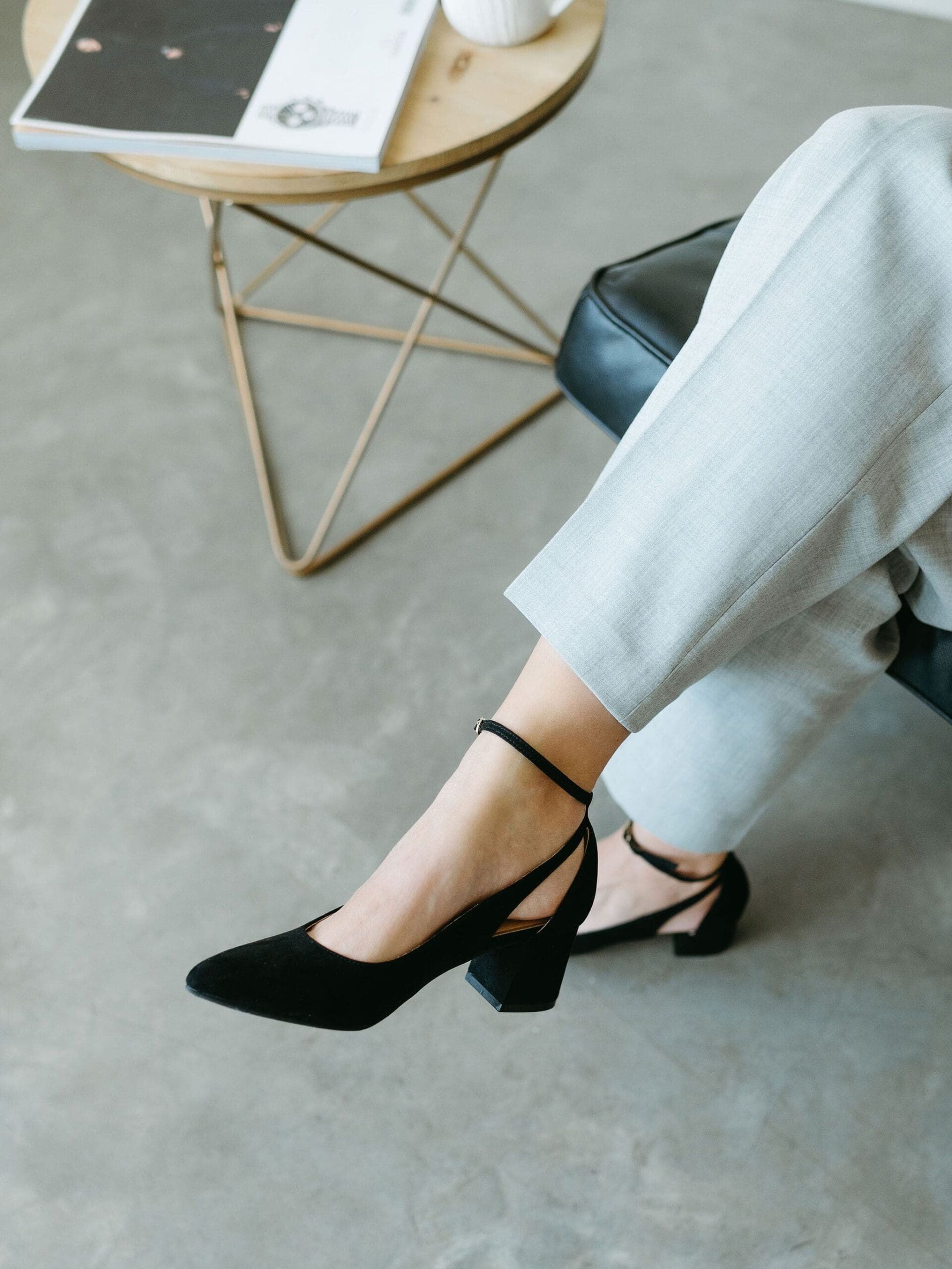 Close-up of a woman wearing black heels and light gray pants, seated near a small side table with a book and coffee mug. | honeybook integrations