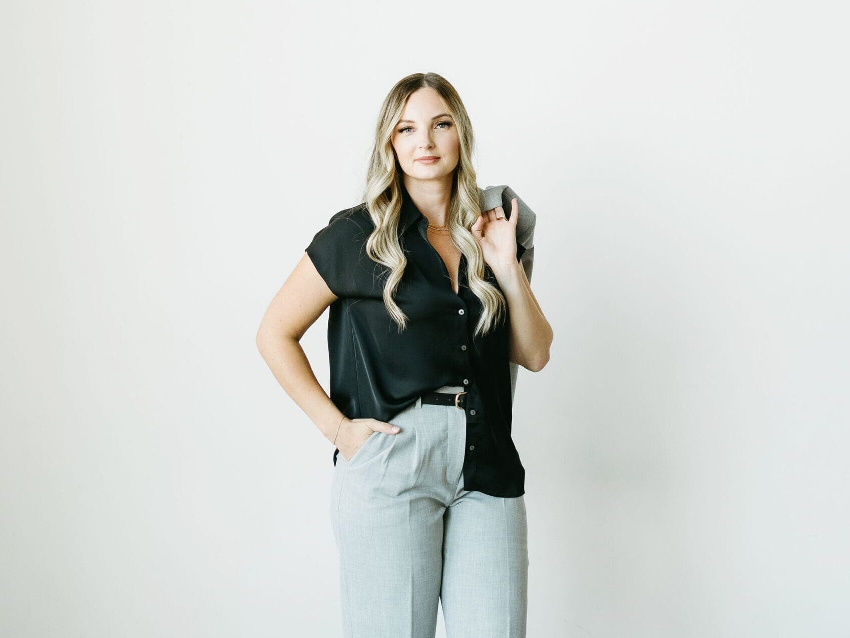 Woman standing against a white wall wearing a black blouse and light gray trousers, styled in a modern professional look. | honeybook integrations