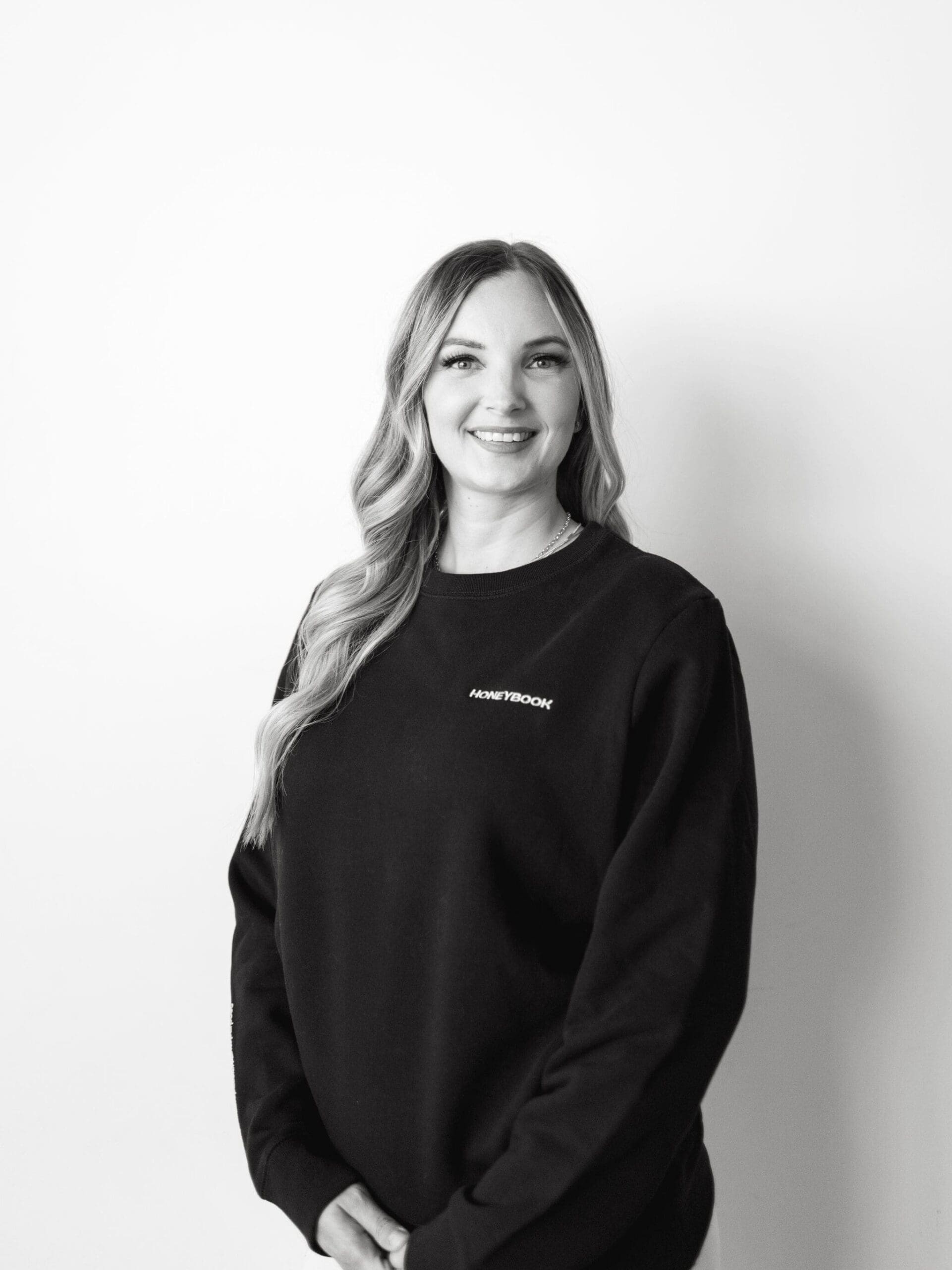 Black and white portrait of a smiling woman wearing a HoneyBook-branded sweatshirt. She stands against a plain white wall, hands relaxed in front of her.