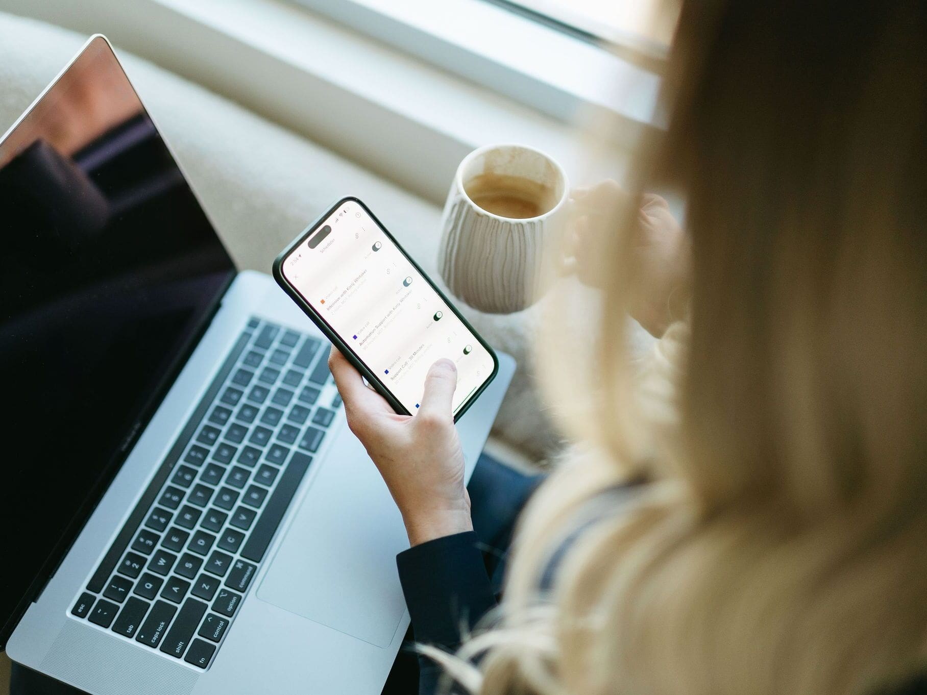 Overhead view of a woman using a smartphone while holding a ceramic coffee mug. A MacBook is open on her lap, and the phone screen shows a list of color-coded tasks or notifications.