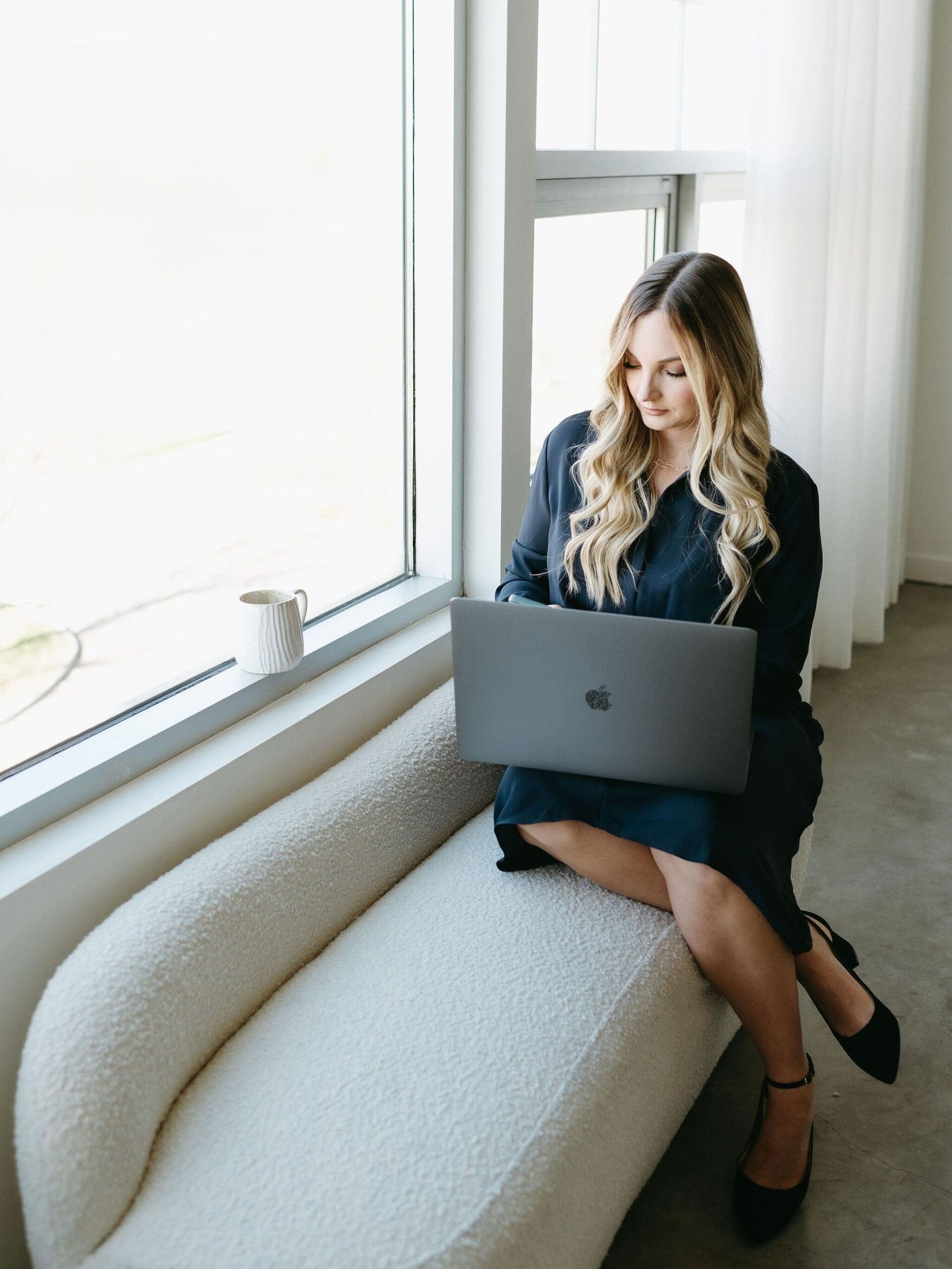 A woman with long blonde hair sits on a cozy cream bench by a large window, working on a MacBook with a coffee mug beside her. Natural light fills the minimalist workspace.