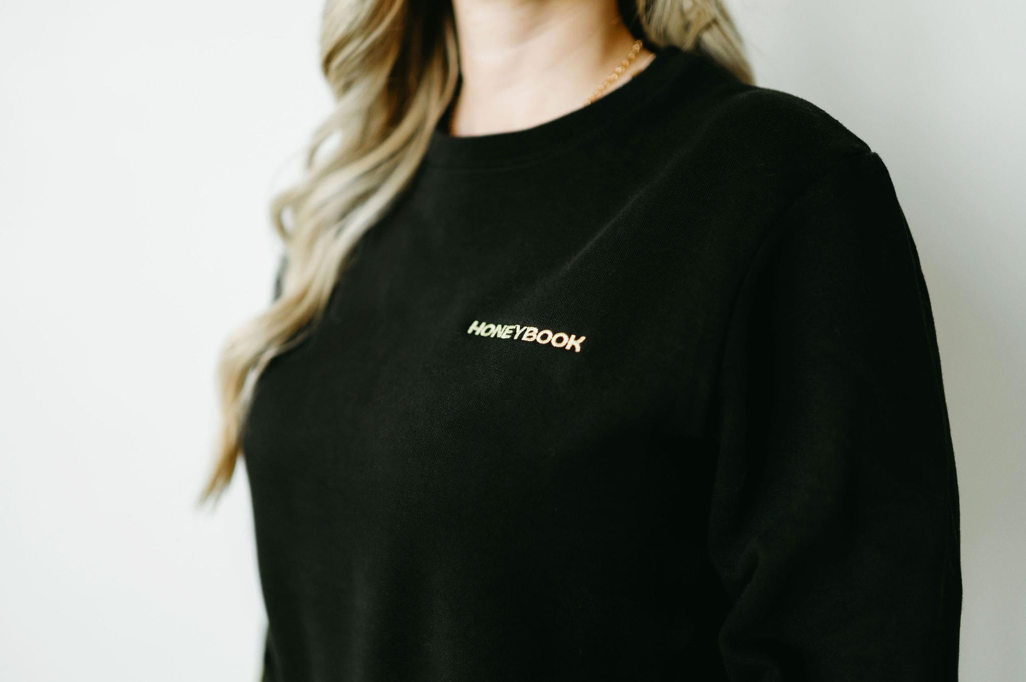 Close-up of a black sweatshirt embroidered with the word “HoneyBook” in gold. The wearer’s long wavy blonde hair is partially visible, along with a gold chain necklace.