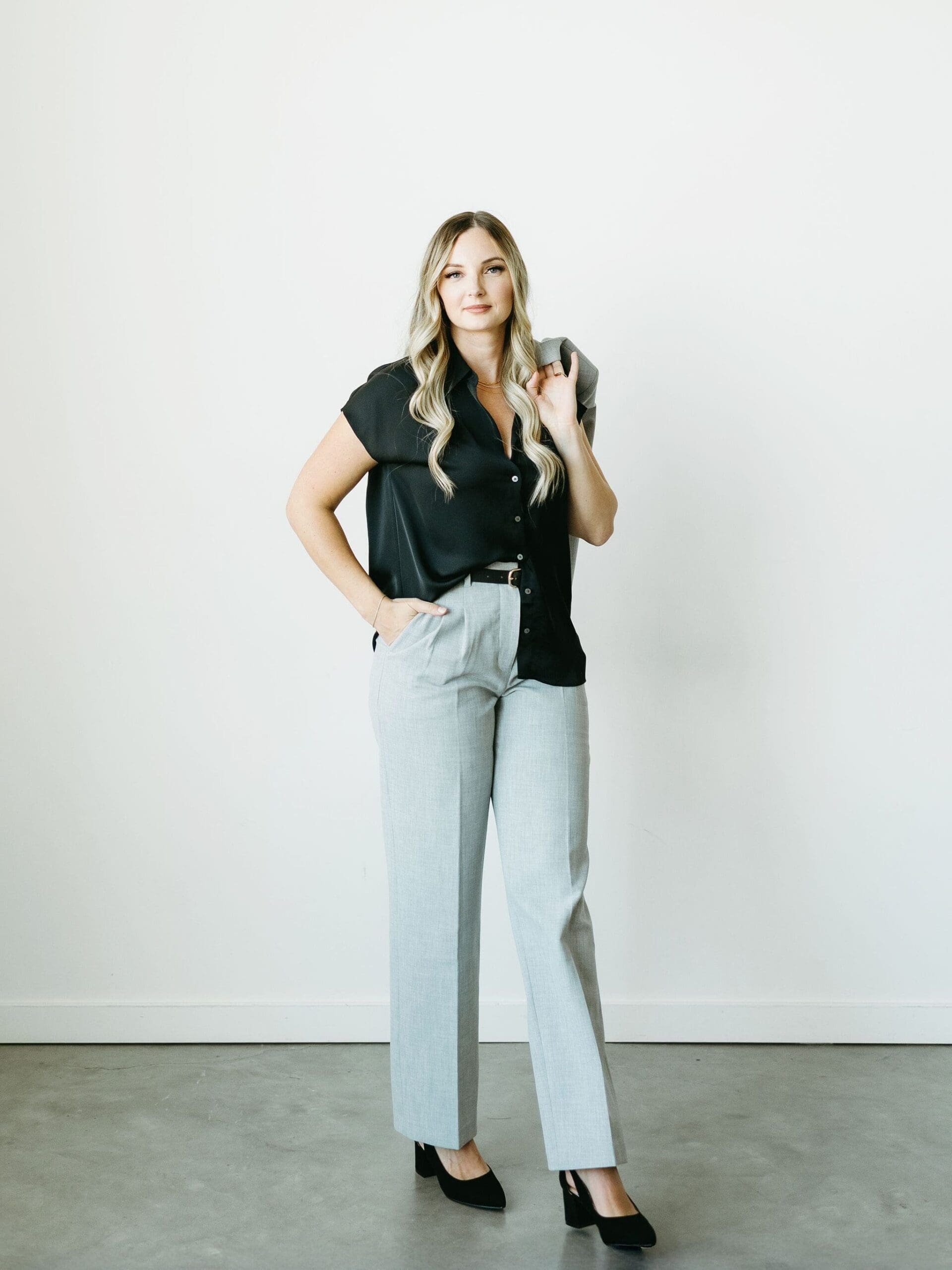 Professional portrait of a female business owner standing against a white wall, dressed in modern business attire, representing a HoneyBook Pro supporting creative entrepreneurs with systems, workflows, and HoneyBook AI.