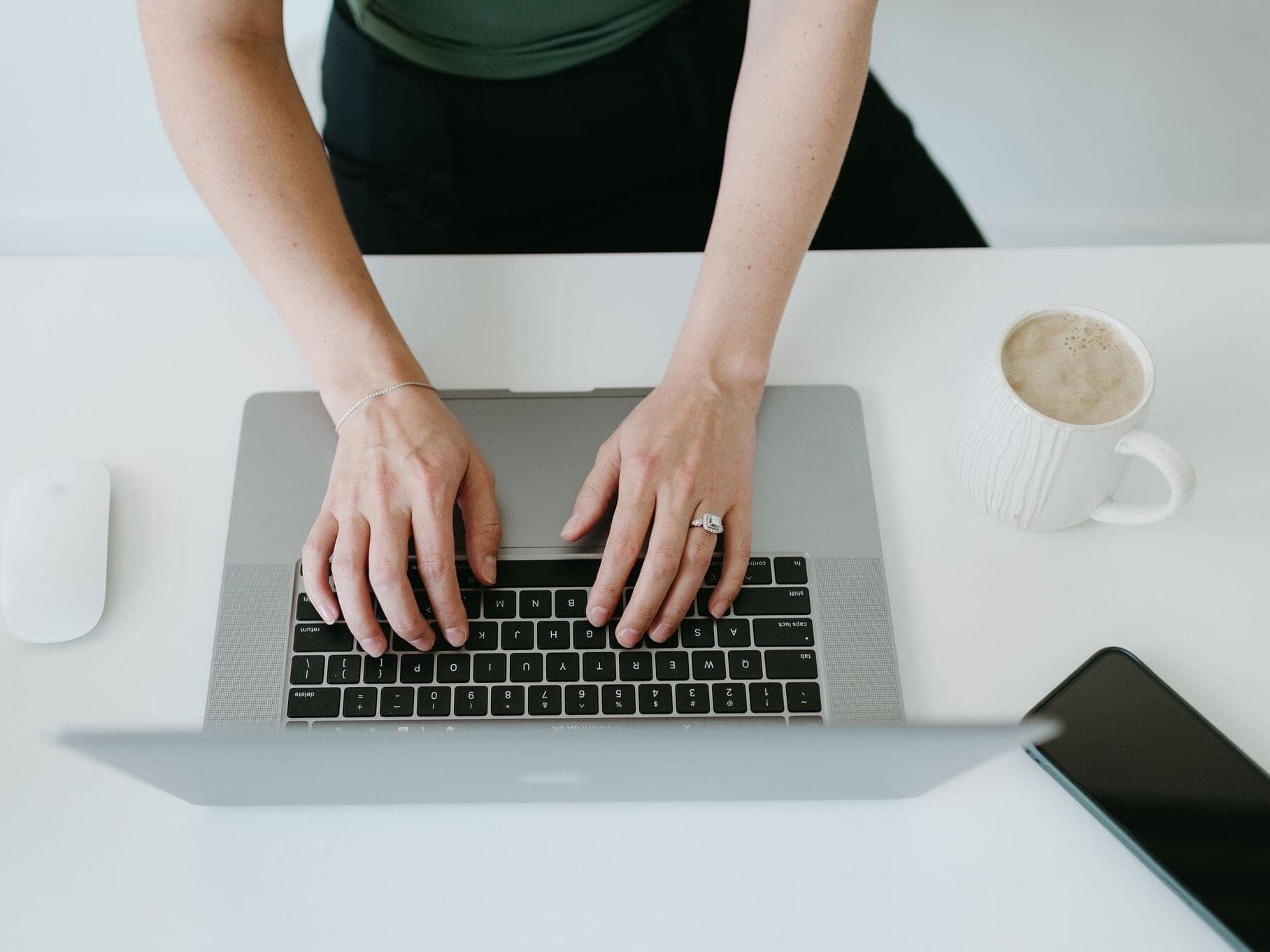 Close-up of hands typing on a laptop at a white desk with coffee and phone nearby, representing a creative entrepreneur managing inquiries, projects, and client communication using HoneyBook AI and CRM workflows.