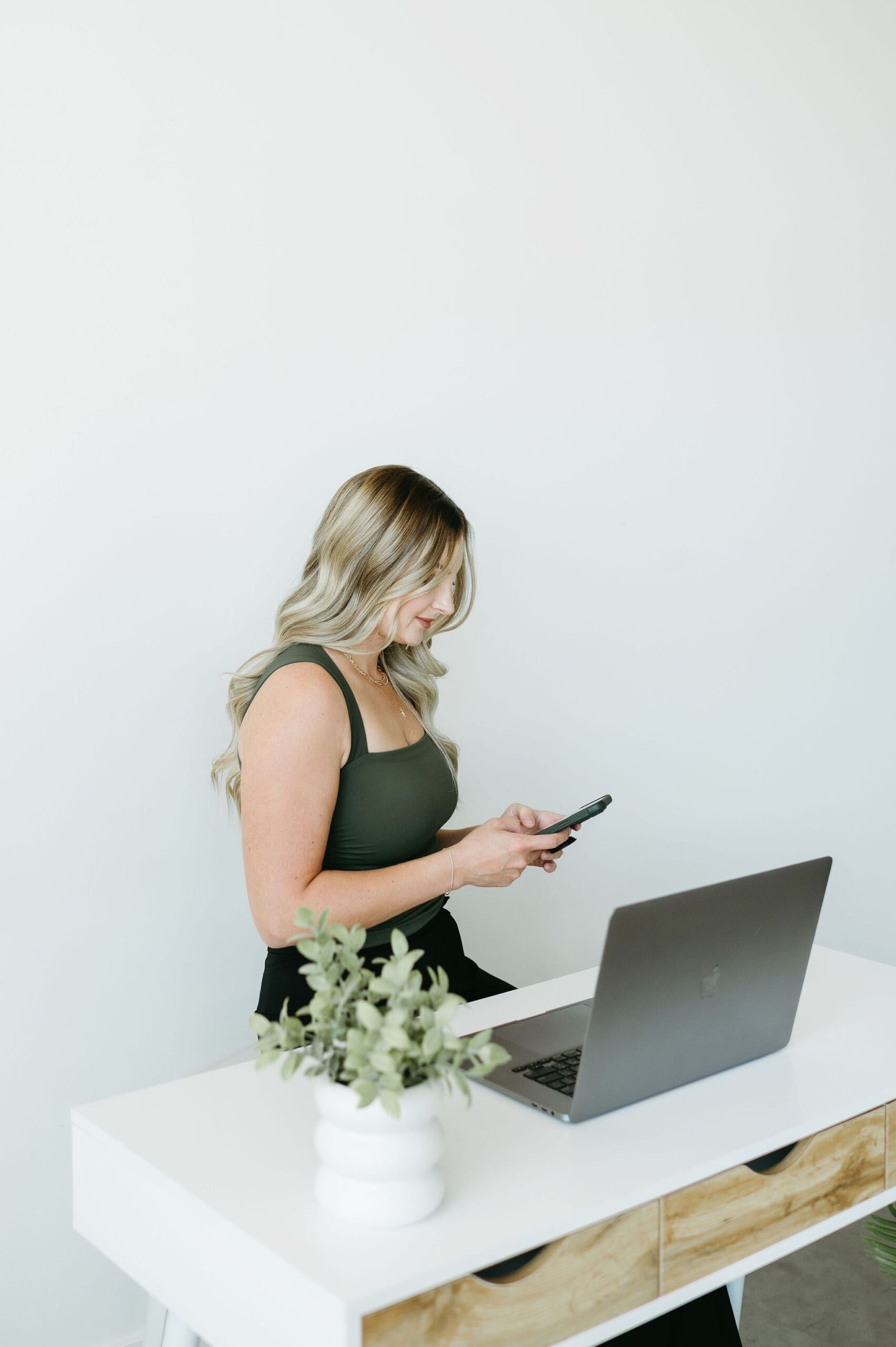 Blonde woman standing at a white desk using her phone beside an open laptop in a minimal office space, representing a creative business owner managing client communication and workflows with HoneyBook AI and CRM tools.