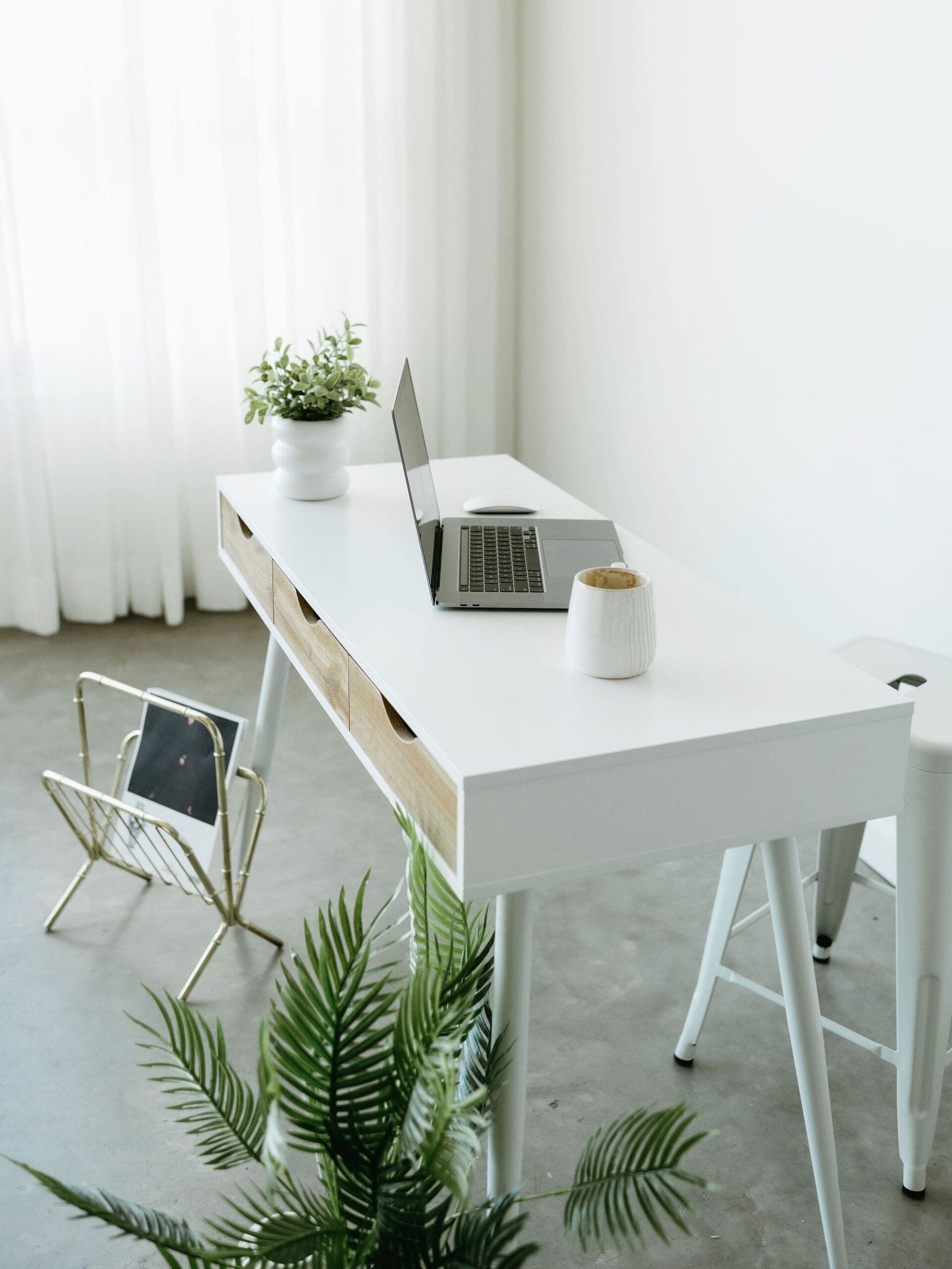 Minimal white home office desk with open laptop, coffee mug, plants, and natural light.