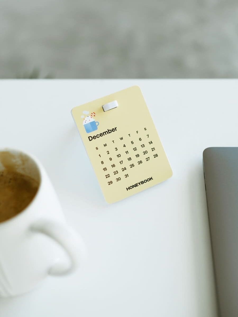HoneyBook branded December calendar card on a white desk beside a coffee cup and laptop, representing scheduling, client management, and CRM organization in a dubsado vs honeybook comparison.