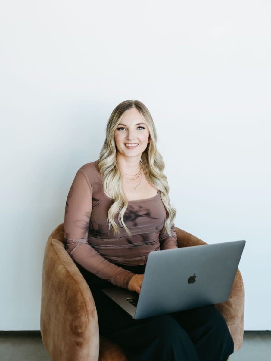 Woman with long blonde hair sitting in a chair working on a laptop, wearing a sheer brown top in a clean, modern workspace with a neutral backdrop.