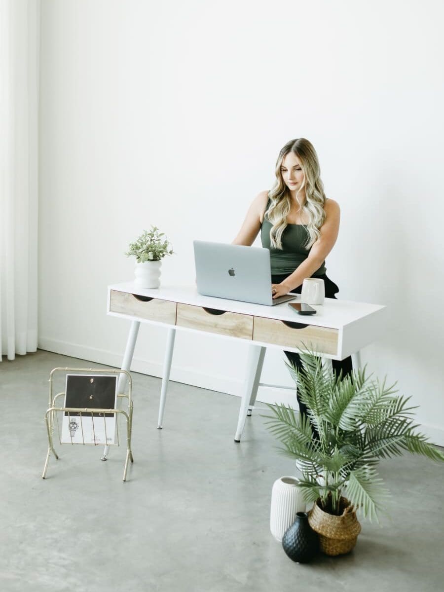 Woman working on a laptop at a white desk in a bright studio, surrounded by plants and minimal decor, creating a calm and organized workspace.