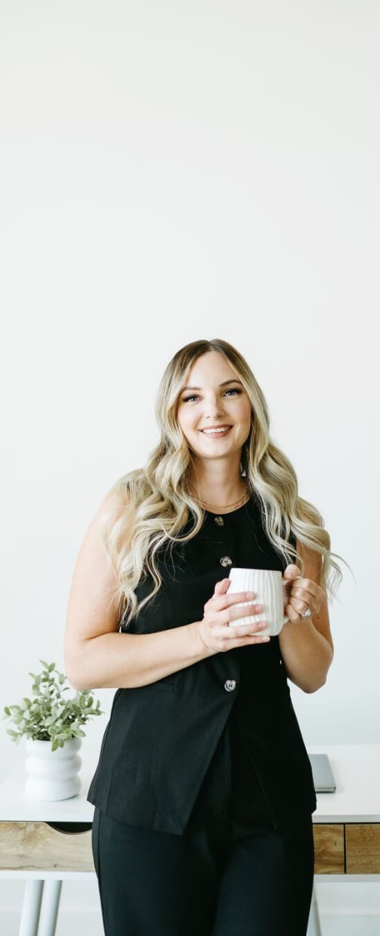 Blonde woman in a black outfit smiling while holding a coffee mug, standing in a minimal office with a white desk, plant, and soft neutral background. HoneyBook pricing
