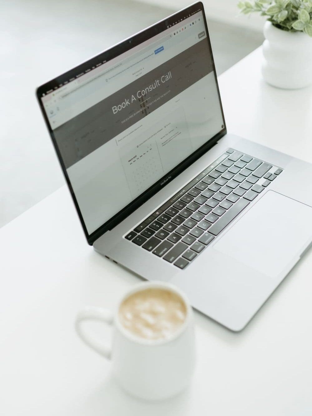 Open laptop on a white desk showing a Book A Consult Call page next to a coffee mug.