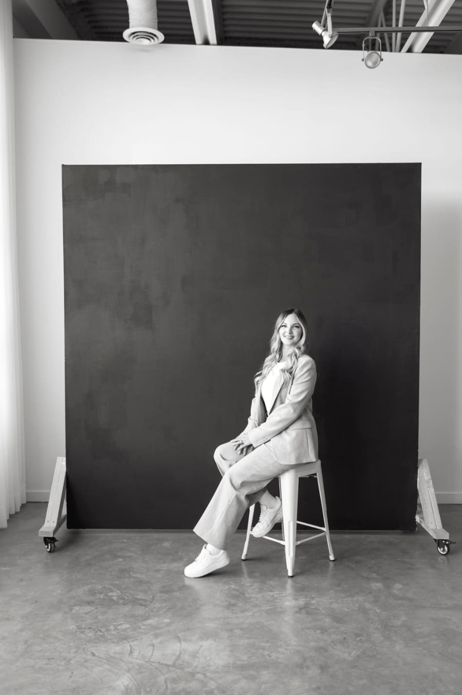 Woman in a neutral suit sitting on a stool in a modern studio, smiling confidently, representing a HoneyBook consultant or creative entrepreneur who has streamlined systems and a polished client experience.