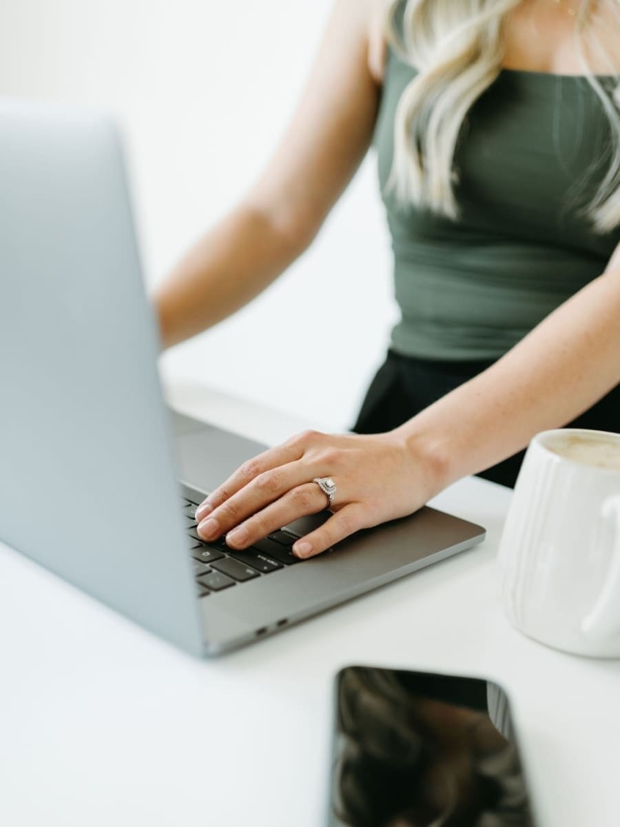 Close-up of hands typing on a laptop with a phone and coffee on a desk, highlighting a business owner working inside their CRM, managing inquiries, automations, and HoneyBook customer service processes.