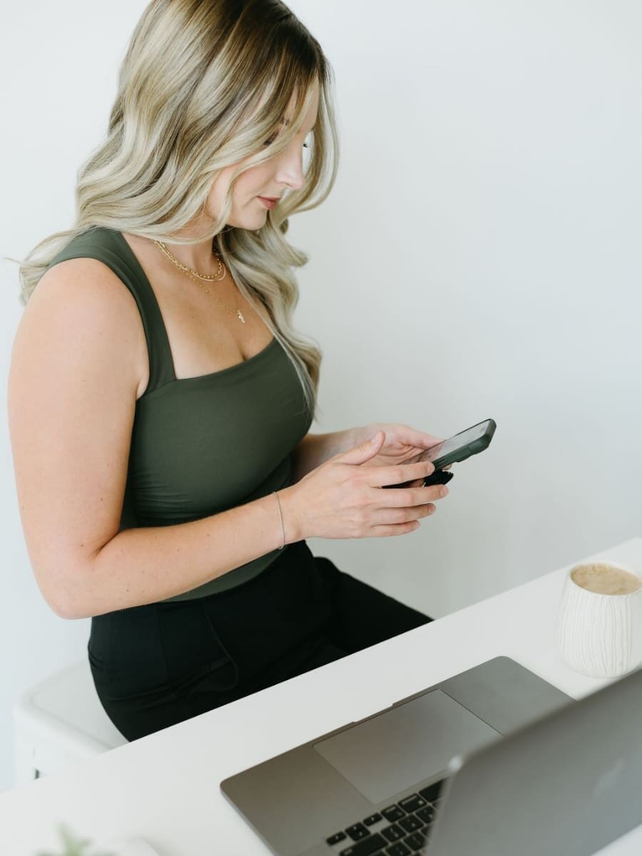 Blonde woman in a green top sitting at a white desk, holding her phone while working on a laptop with a coffee nearby, representing a creative entrepreneur managing client communication and HoneyBook customer service tasks.