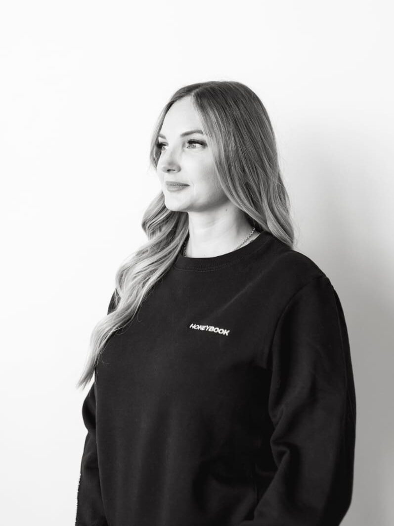 A black-and-white portrait of a woman wearing a HoneyBook sweatshirt, looking confidently ahead. The minimalist composition conveys professionalism and brand pride, representing creative entrepreneurs who rely on HoneyBook workflows.