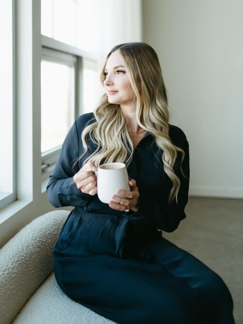 A woman with long blonde hair sits by a bright window holding a white mug, dressed in a navy outfit. The calm, professional setting reflects focus and balance—perfect for illustrating productivity and streamlined HoneyBook workflows.