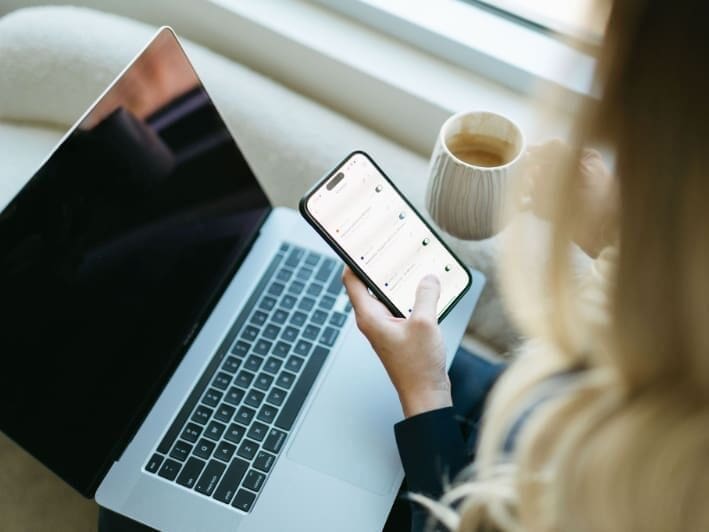A close-up of a workspace showing a laptop, smartphone, and coffee mug. The person reviews messages on their phone, symbolizing modern business efficiency and how HoneyBook workflows help manage client communication seamlessly.