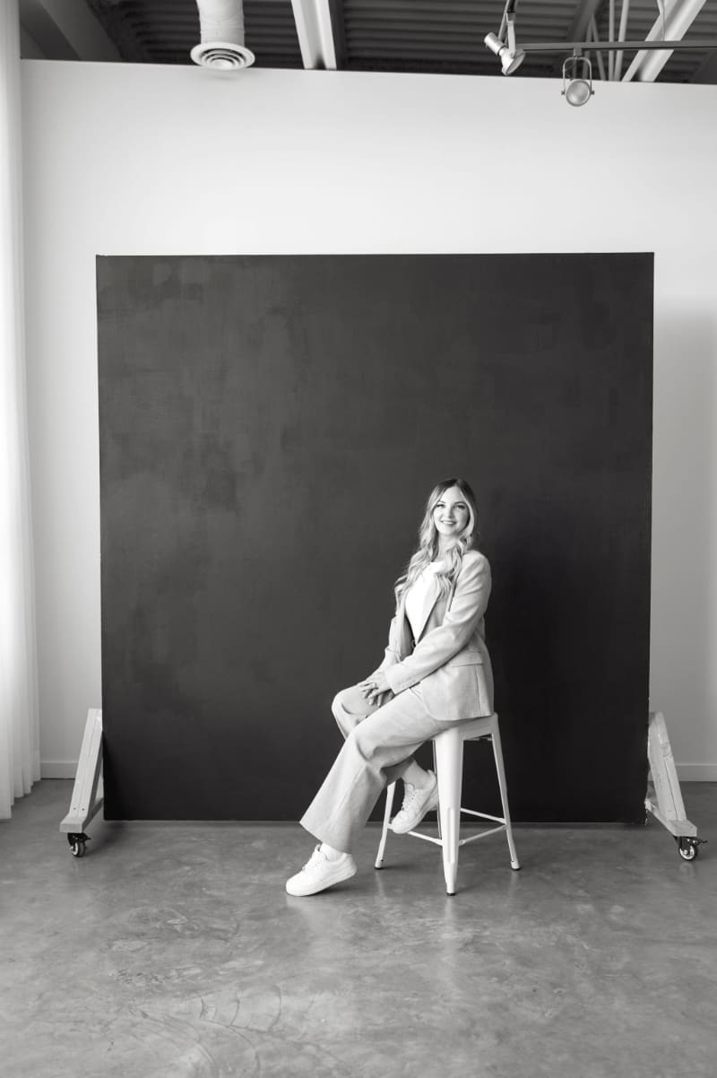 Karly Whitaker, HoneyBook consultant, sitting on a stool against a black backdrop, smiling in a tan suit. Helping creative entrepreneurs set up HoneyBook and QuickBooks systems for a smoother backend.