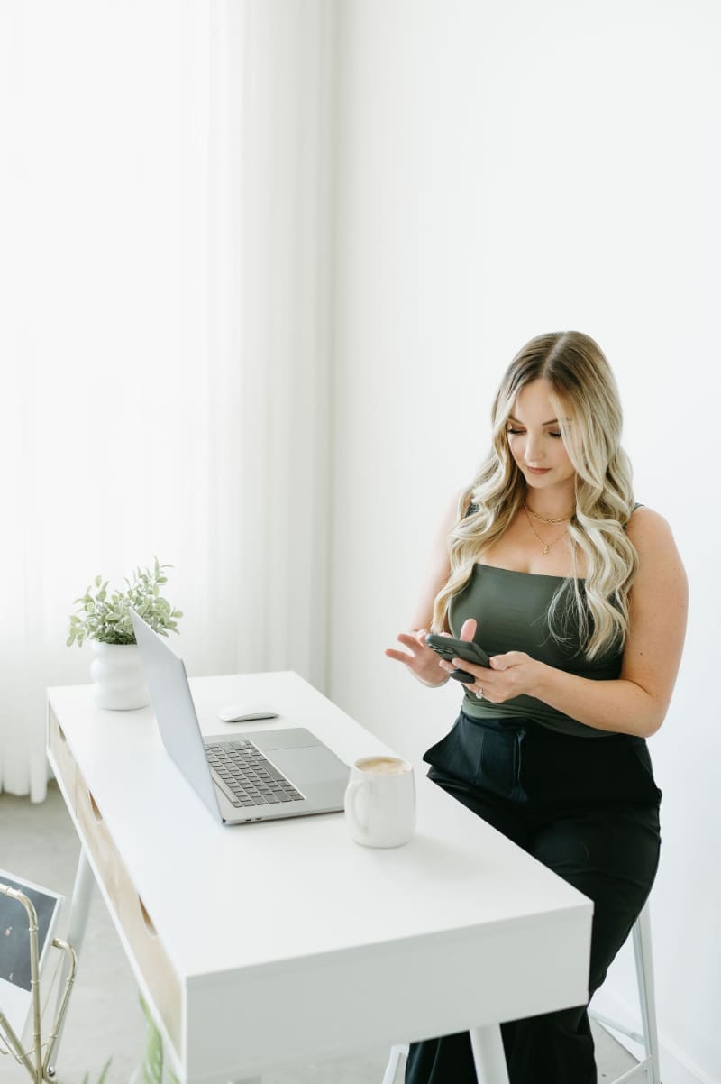 Karly Whitaker planning what to set up first in HoneyBook, phone in hand at a white desk with an open laptop and coffee.