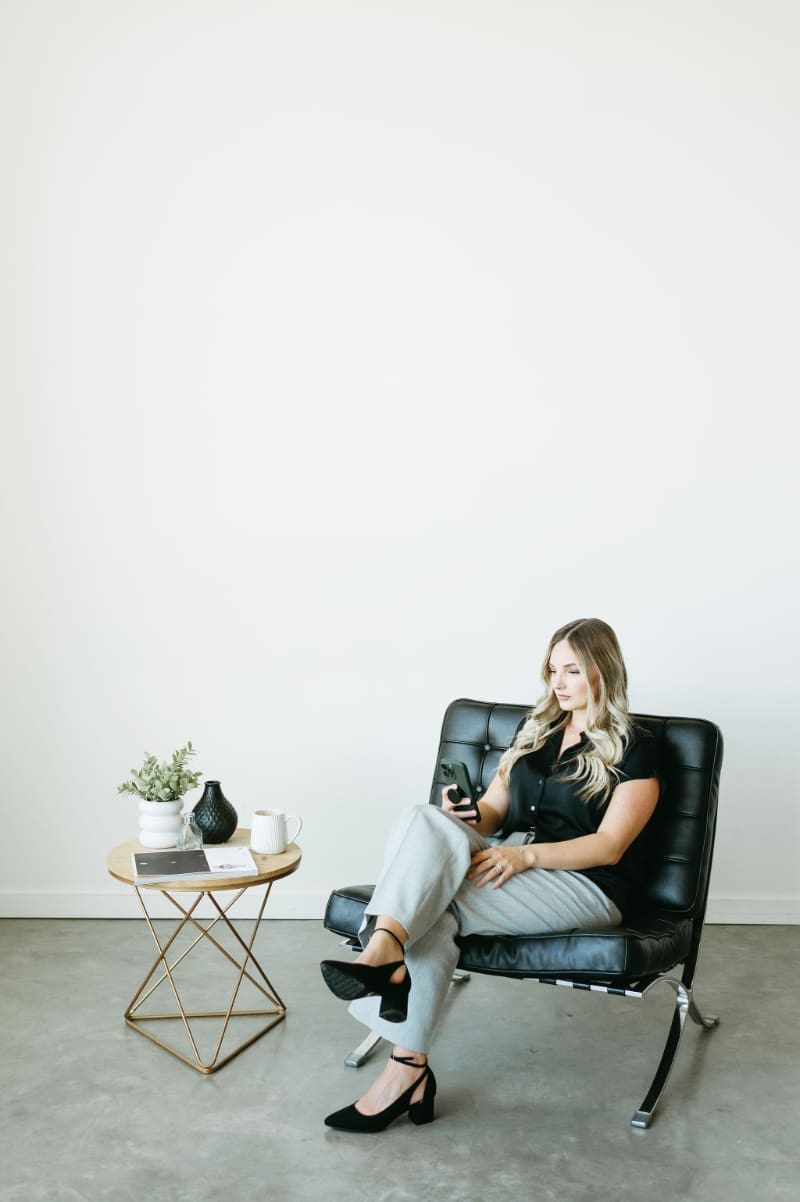 HoneyBook consultant Karly Whitaker seated in a black leather chair reviewing her phone beside a gold side table and coffee.