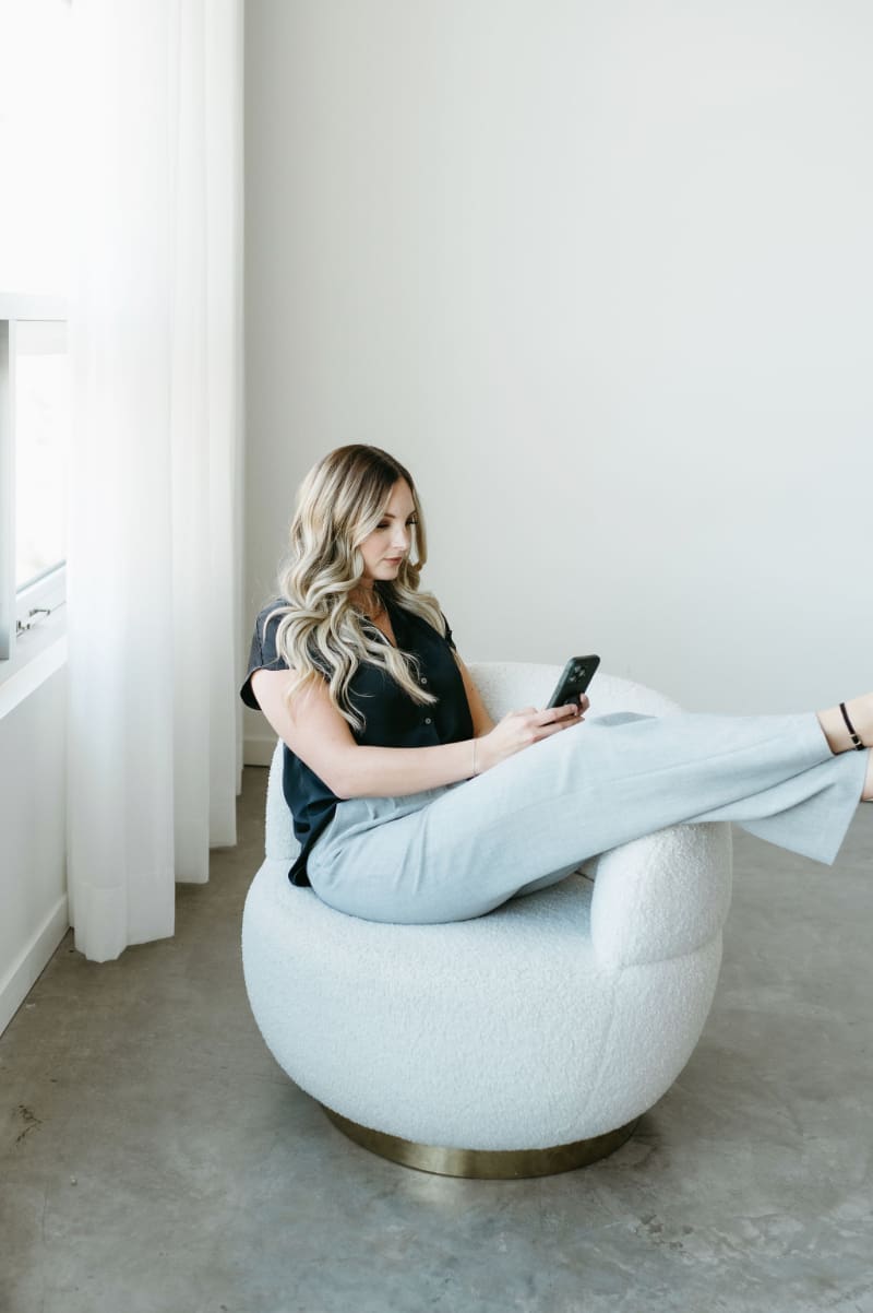 Karly Whitaker relaxing sideways in a white boucle chair with her phone, showing what a calm HoneyBook workflow looks like.