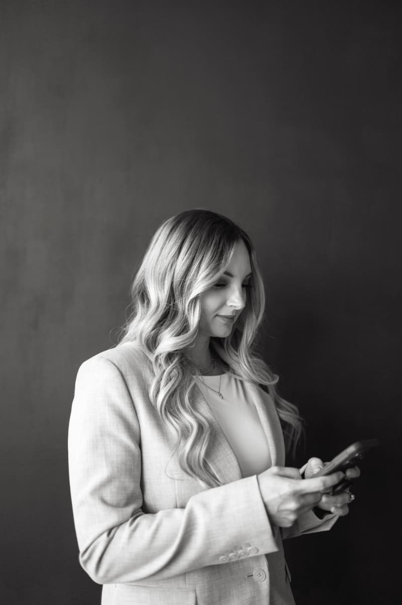 Black-and-white portrait of a business owner checking her phone while wearing a blazer, reflecting the communication and client follow-up that can happen after sending a HoneyBook proposal.