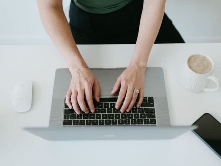 Overhead view of a business owner typing on a laptop at a white desk with coffee, phone, and mouse nearby, representing the process of creating and customizing a HoneyBook proposal for a client.