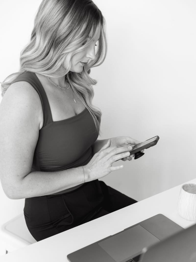 Business owner checking her phone beside her laptop, showing how the HoneyBook scheduler helps simplify client scheduling and reduce back-and-forth booking emails.