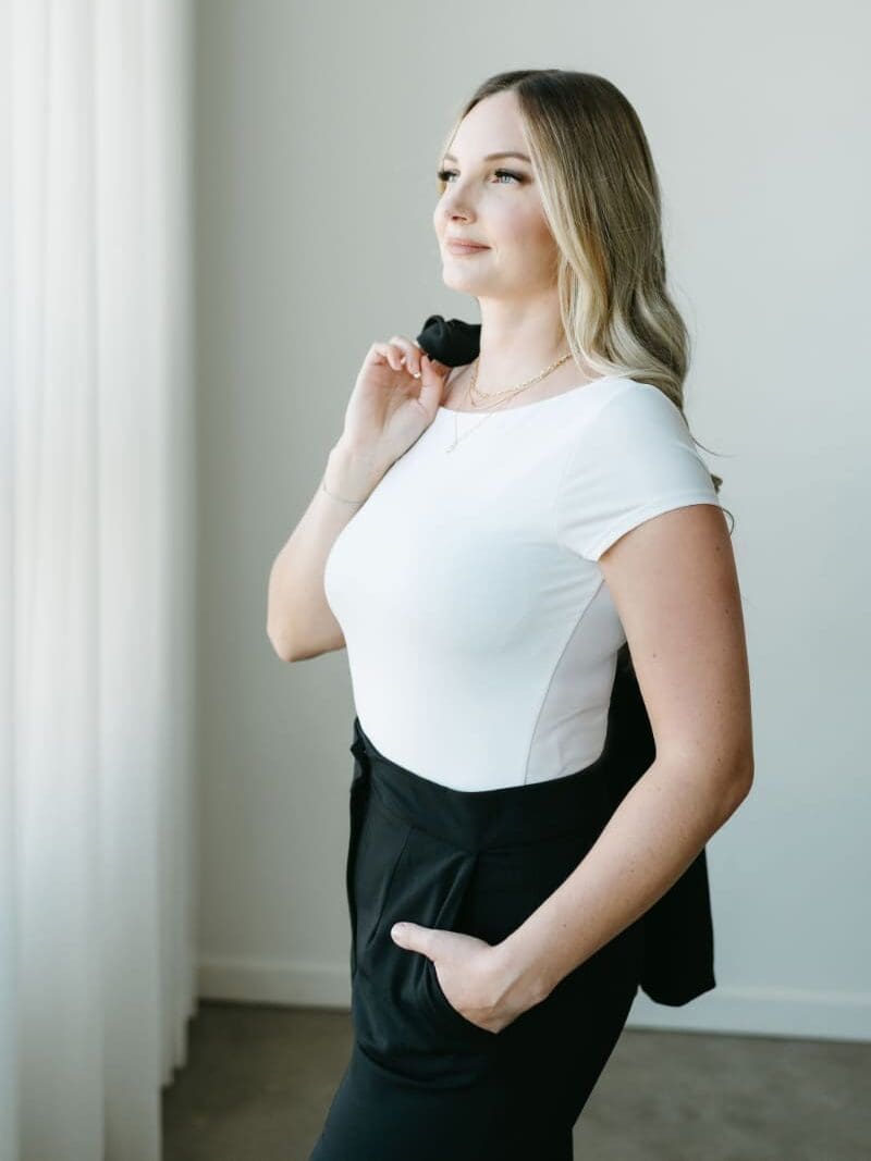 Professional branding portrait of HoneyBook Pro Karly Whitaker standing beside a bright studio window, wearing a white top and black trousers with a jacket over her shoulder. Featured in her tutorial on multiple project dates in HoneyBook for service providers.
