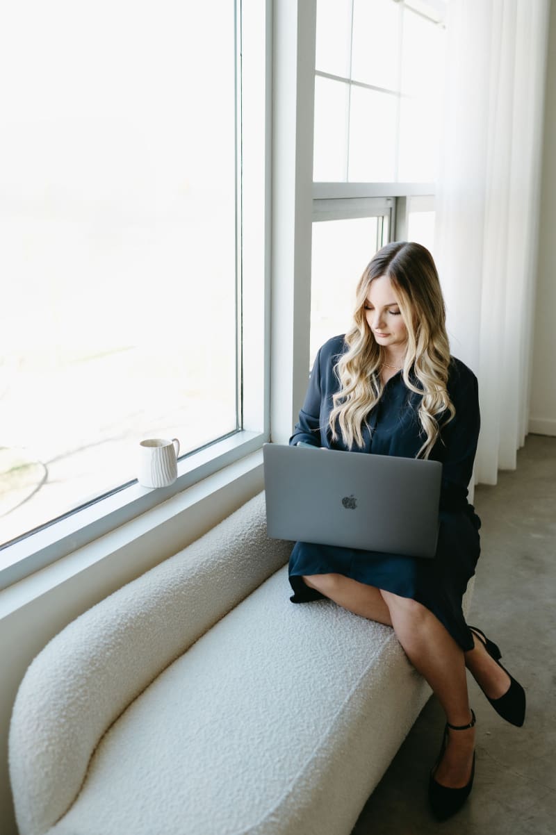 HoneyBook expert Karly Whitaker sitting on a cream window bench with a laptop, working in a bright studio with large windows and a coffee mug nearby. A behind-the-scenes branding photo for her guide to managing multiple project dates in HoneyBook and client workflows.