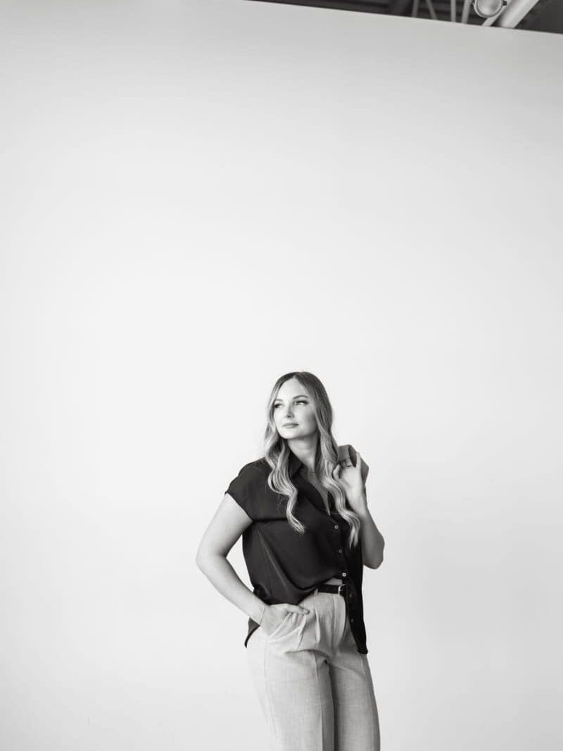 Black-and-white branding portrait of HoneyBook pro Karly Whitaker standing against a white studio wall, wearing a dark blouse and light trousers. A professional headshot for her guide to multiple project dates in HoneyBook and managing client bookings.